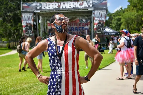 Man wearing a stars and stripes sleeveless shirt has his hands on his hips while standing in front of a music stage with a large Beer 'Merica banner at the top.