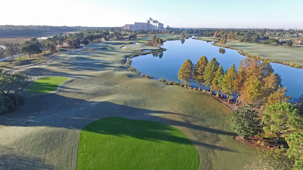 Aerial view of course hole with the green at the front, a curved, hilly fairway that runs alongside a large pond. In the background is a large resort-type building.