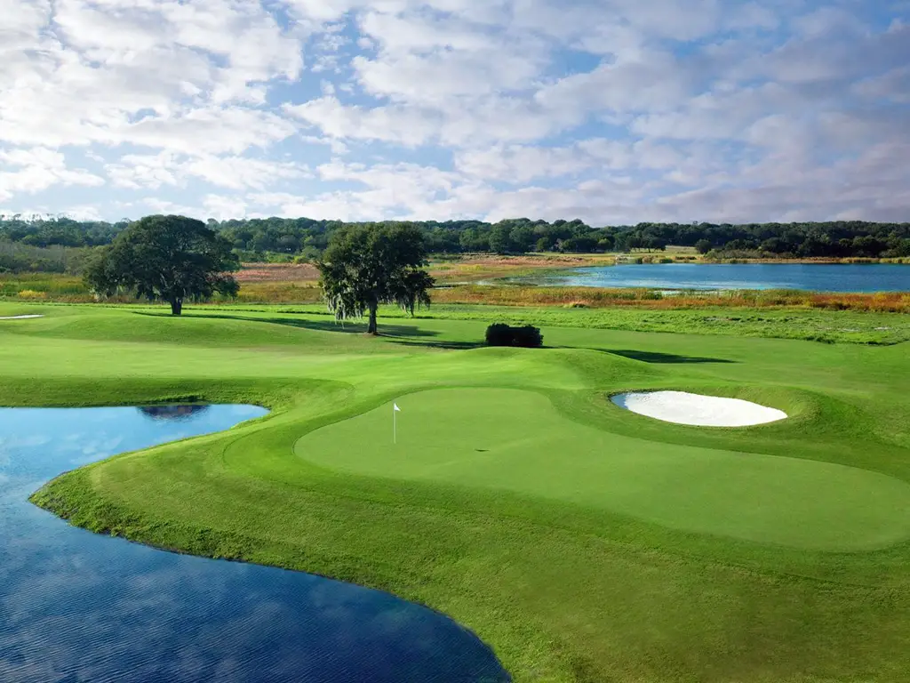 Golf course hole with green in foreground with pond on left side and small sand trap on right side. A few trees line the fairway.